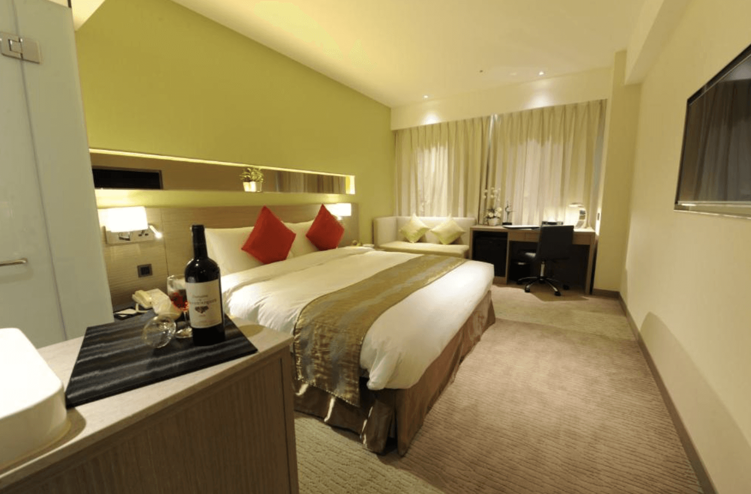 【小港住宿】高雄國際會館的客房，房內以暖色系為主，設有寬敞的雙人床，床上擺放著紅色抱枕，旁邊有沙發、書桌與辦公椅，適合商務人士或情侶入住。房間內光線柔和，氣氛溫馨放鬆，前景則有一瓶紅酒與杯子。