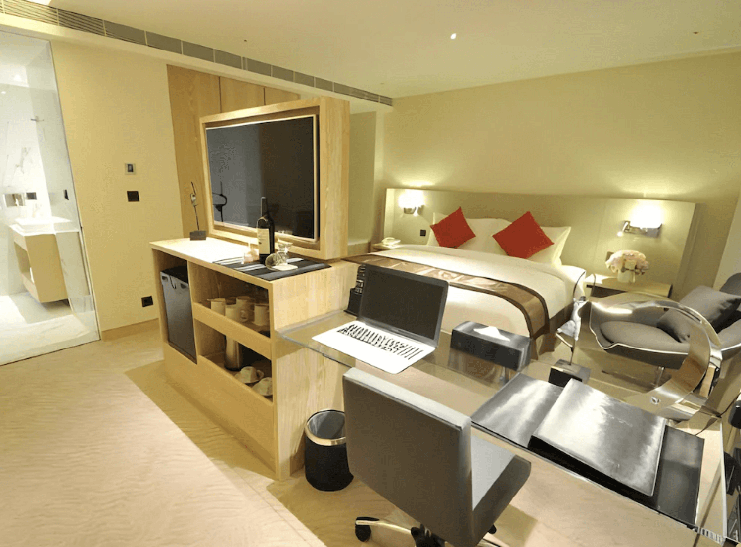 【小港住宿】高雄國際會館的房間內部設計，設有透明玻璃工作桌，配備筆記型電腦與椅子，十分適合商務旅客。中央有木質隔間櫃，上方放置大型液晶電視與茶水區，背景是舒適的大床與床頭櫃，空間格局現代且實用。