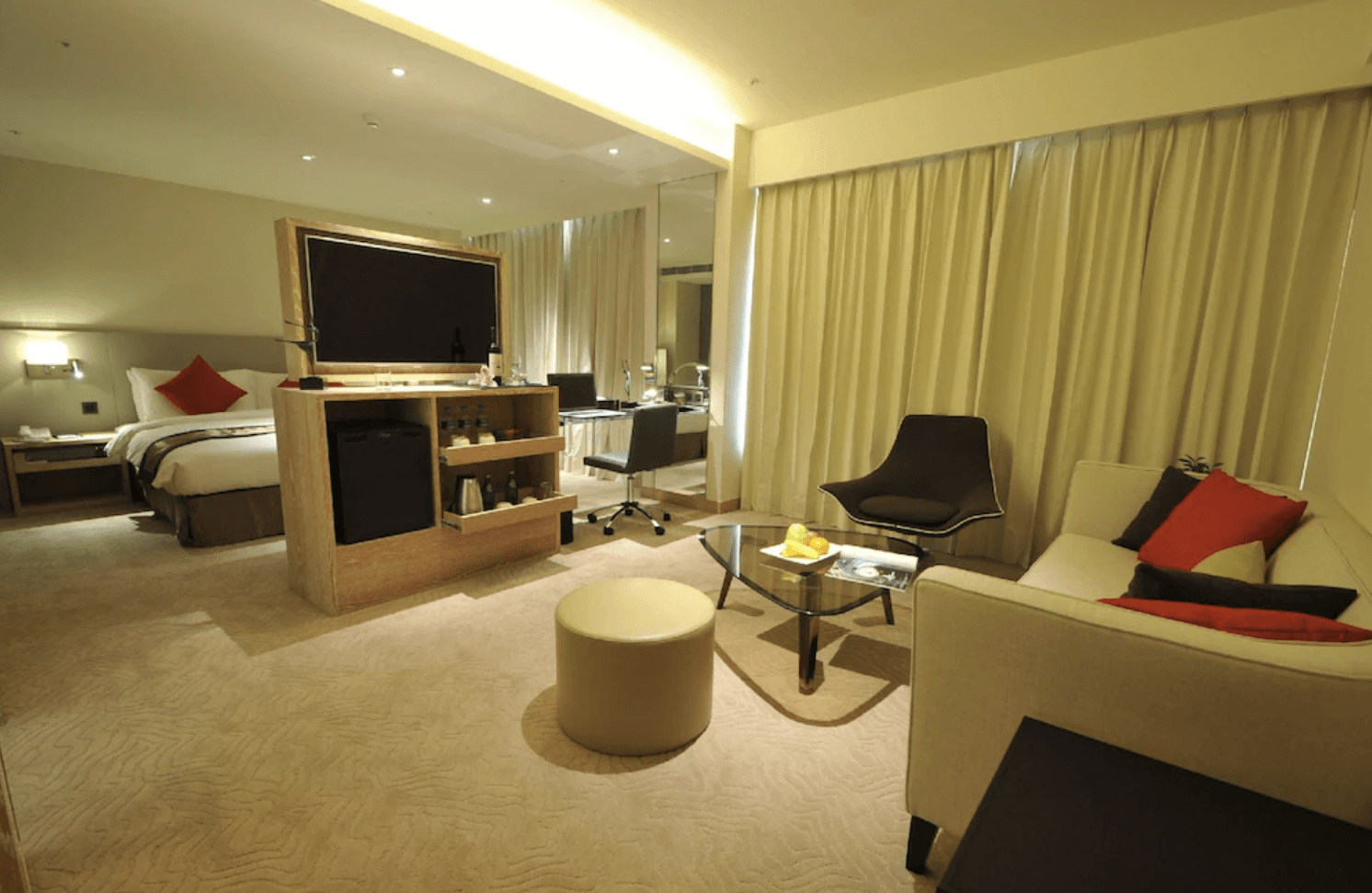 【小港住宿】高雄國際會館的套房，整體空間非常開闊，有明亮的客廳區域，包括舒適的沙發、茶几、現代感座椅及圓形椅凳，配有精緻的小吧台和液晶電視，後方則是床鋪區域，整體色調溫暖柔和，適合放鬆休閒或小型會客。