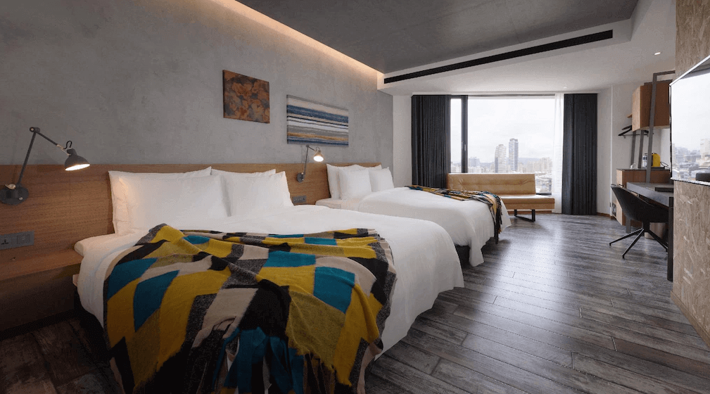 【高雄親子飯店】“高雄中央公園住宿”高雄比歐緻居的雙床房，房間內採用簡約工業風格，牆面為灰色混凝土質感，搭配溫暖木質床頭板與內嵌燈帶。房內擁有大片落地窗，可欣賞高樓都市景觀，並設有長型沙發座椅。床鋪上鋪有幾何圖案的毛毯，增添設計感。