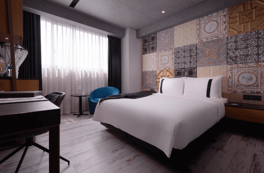 【高雄親子飯店】高雄比歐緻居的特色雙人房，牆面裝飾有復古磁磚拼貼圖案，營造歐式懷舊風格。房內配備一張舒適的大床，床頭燈採用現代工業風設計，並有一張藍色單椅與小茶几，提供休閒空間。窗戶搭配白色紗簾與黑色厚窗簾，柔和自然光線進入房內。