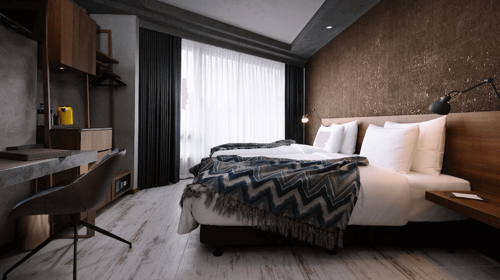 【高雄親子飯店】高雄比歐緻居的溫暖木質調雙床房，房內採用深色紋理牆面與木製家具，展現沉穩的工業風設計。床頭燈為可調式壁燈，適合閱讀使用。房內設有辦公桌與置物架，並提供膠囊咖啡機，提升住宿便利性。