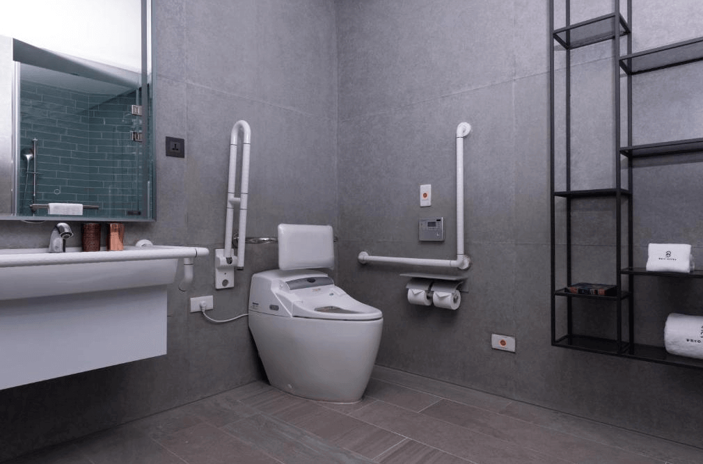 【高雄親子飯店】高雄比歐緻居的無障礙衛浴空間，整體以灰色系磁磚為主，搭配簡約黑色金屬置物架。浴室內配有無障礙輔助設施，包括安全扶手、符合人體工學的坐式馬桶，以及方便輪椅使用者的懸空洗手台設計，展現貼心的無障礙設計。