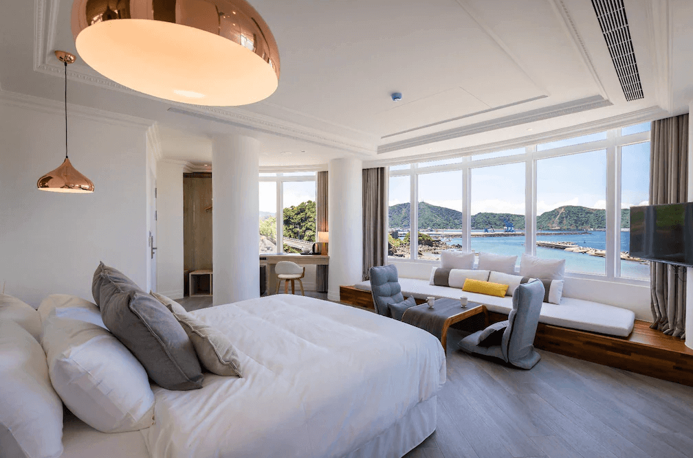 【蘇澳住宿】BRIGHTHOUSE 光之旅的海景雙人房，設有一張大床、舒適的沙發區與落地窗，可遠眺港灣與山景，室內採白色與木質設計，營造出溫馨且現代的氛圍。