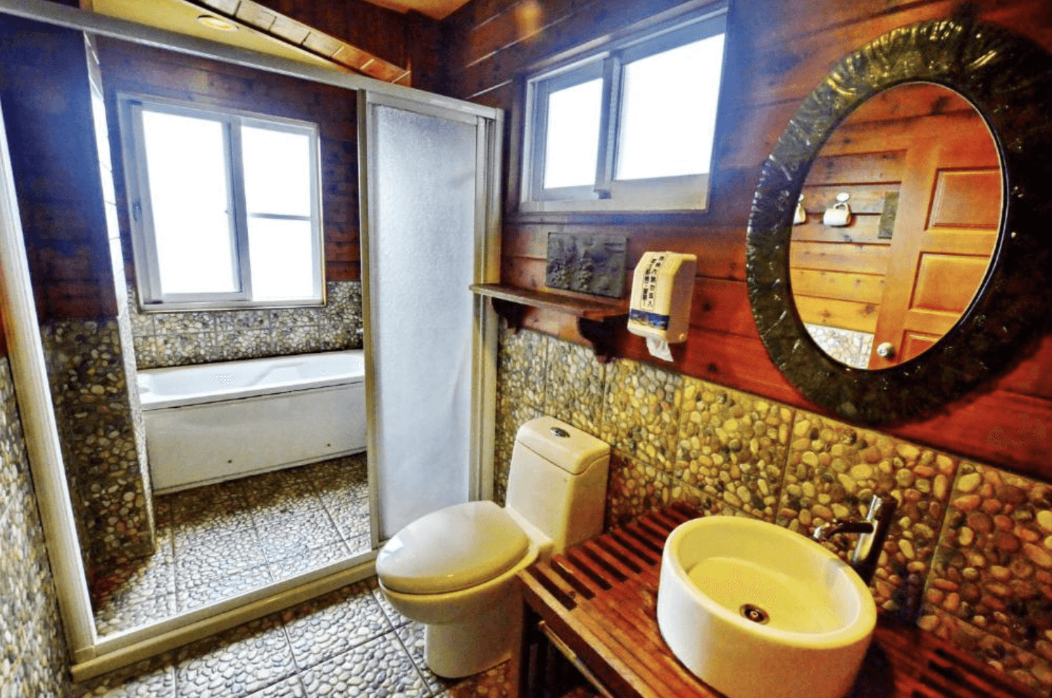 【清境農場住宿】事外桃源渡假山莊的山林風格的浴室，牆面與地面為鵝卵石設計，配備馬桶、洗手台與乾濕分離淋浴間，並設有浴缸與對外窗戶，自然採光充足，營造放鬆舒適的沐浴空間。