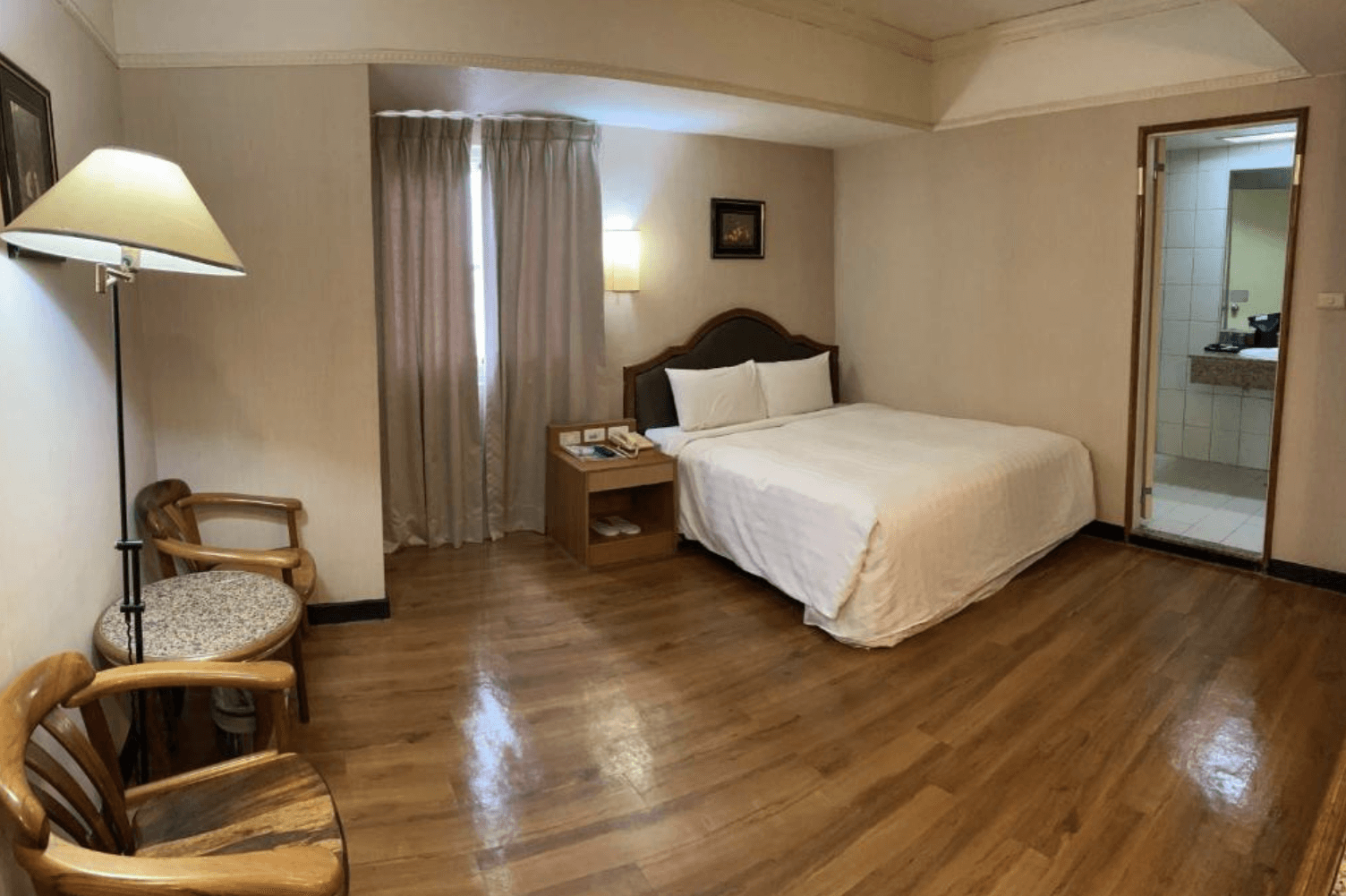 【東港住宿】大鵬灣大飯店的雙人房，設有一張雙人床與木質地板，搭配兩張木椅與小茶几，採用柔和燈光與窗簾營造舒適氛圍，房內可見獨立衛浴空間。