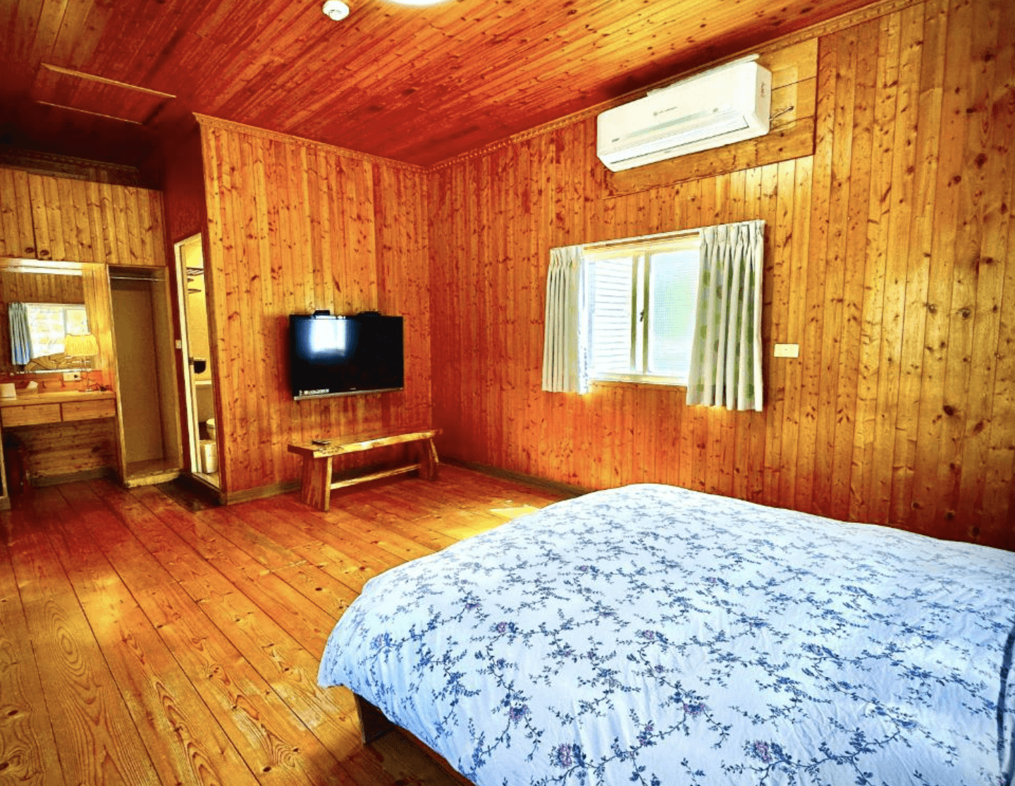 【三義住宿】峯州小木屋簡約寬敞的雙人房，全室鋪設木地板與木牆，牆上配有壁掛電視與冷氣，窗邊設有梳妝區，展現自然溫潤的空間感受。
