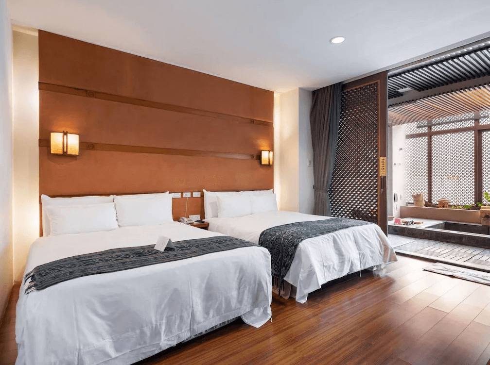 【谷關住宿】惠來谷關溫泉會館的房間內設有兩張雙人床，搭配木地板與溫潤橘色牆面，散發自然溫暖的住宿氛圍。房內可直通半露天泡湯空間，拉門設計保有隱私又能通風，適合家庭或朋友一同放鬆住宿。