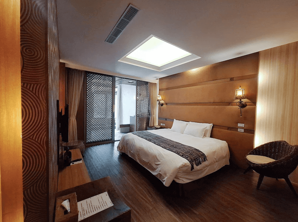 【谷關住宿】惠來谷關溫泉會館木質調與柔和燈光交織的雙人房，床頭設有古銅色壁燈與民族風掛飾，營造沉靜的異國風情。大片玻璃拉門通往半戶外泡湯空間，房內設有天井採光，白天自然光充足。