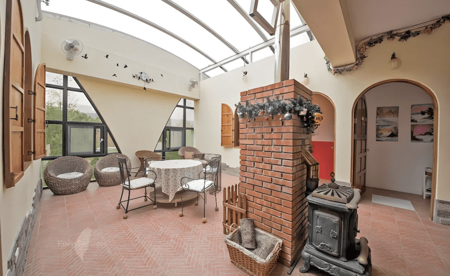 【三義住宿】慕哲哈比屋的室內陽光玻璃屋空間，配有壁爐與紅磚牆，擺設典雅的圓桌與藤椅，營造溫馨鄉村氛圍，適合休憩與交流。