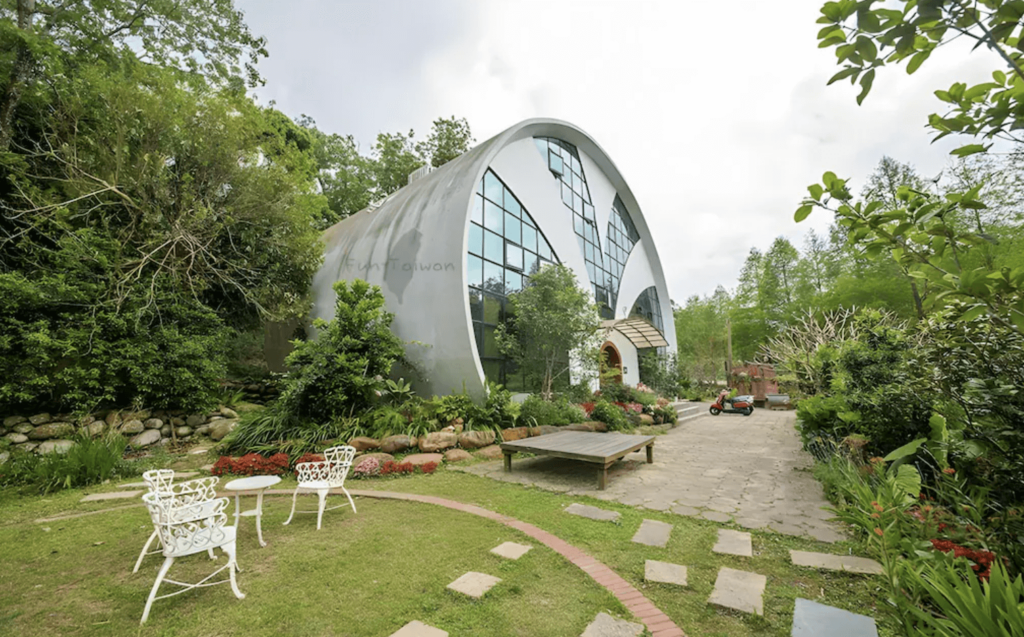 【三義住宿】"三義民宿精選圖示"慕哲哈比屋的獨特拱形建築外觀，位於綠意盎然的庭園中，前方擺有白色歐式戶外桌椅，環境清幽，彷彿童話世界中的小屋。