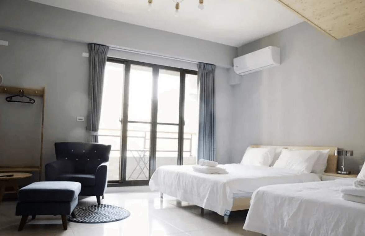 【東港住宿】東帆巷弄休閒民宿的房內設有兩張雙人床，採光良好，大片落地窗搭配灰藍色窗簾，配備冷氣、單人沙發與腳凳，整體色調柔和，營造出舒適又放鬆的住宿氛圍。
