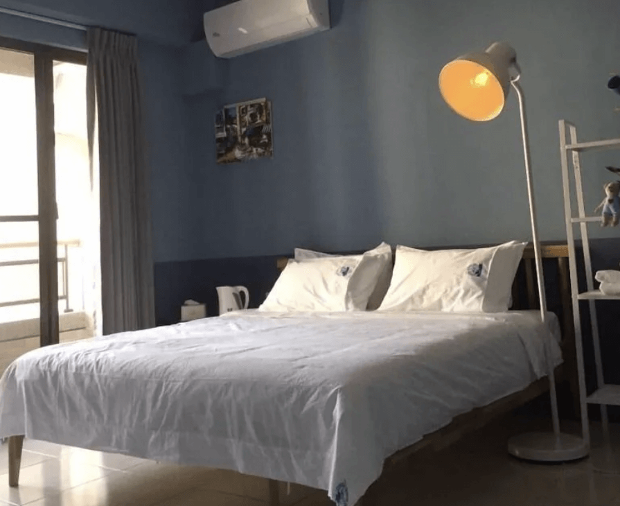 【東港住宿】東帆巷弄休閒民宿的藍色牆面搭配溫暖黃光的立燈，雙人床鋪上白色寢具，床旁設有熱水壺與簡易收納架，整體風格溫馨沉靜，適合情侶或單人入住。