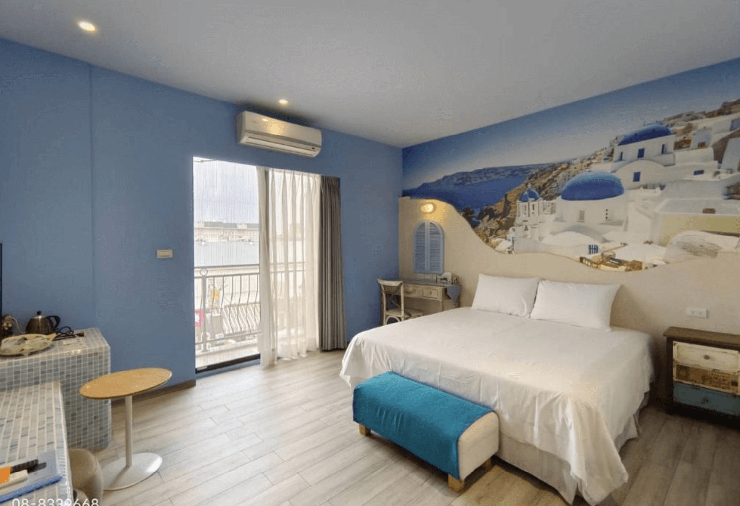 【東港住宿】東港幸福168民宿的房間牆面繪有希臘聖托里尼藍白小屋壁畫，搭配淡藍色牆面與木紋地板，設有一張雙人床、白色書桌、藍色腳凳與大面窗簾門，營造出地中海度假氛圍。