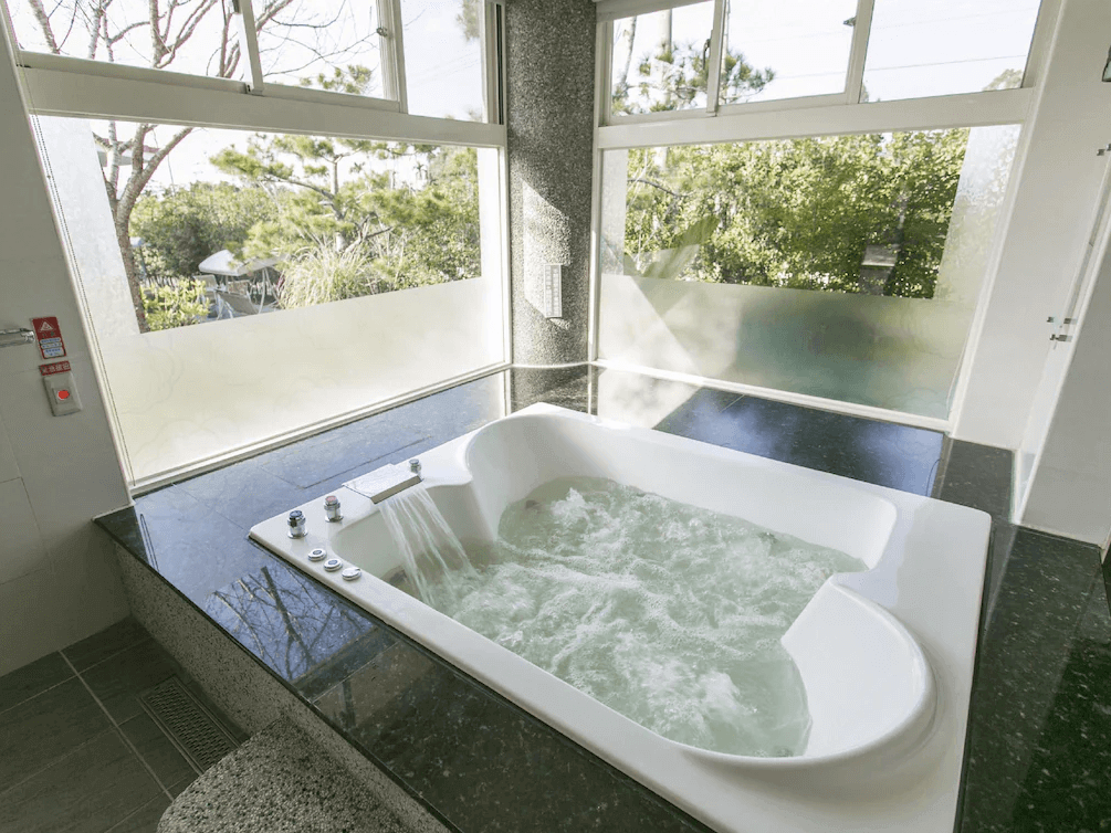 【關子嶺溫泉】“白河住宿”林桂園石泉會館擁有大片落地窗的室內景觀湯池，搭配白色按摩浴缸與流水設施，窗外可見綠意盎然的自然景致。