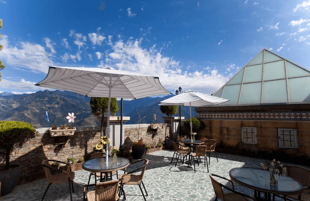 【清境農場住宿】清境夏都夢幻山林渡假山莊在山林中的露天觀景平台，設有多組圓桌與藤椅，搭配大型白色陽傘，背景是藍天白雲與群山景致，是享受午後陽光與山景的理想地點。