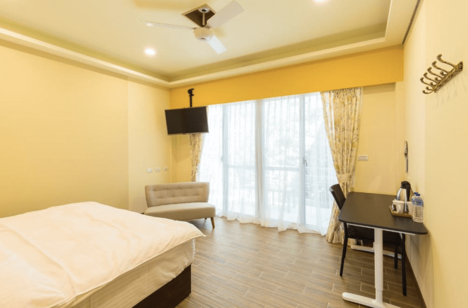 【清境農場住宿】清境繽紛農場的雙人房，牆面以淡黃色為主色，搭配木質地板與簡約家具，包括一張雙人床、灰色小沙發、壁掛電視與工作桌，大片落地窗透入自然光，窗簾為碎花圖案，整體風格溫暖舒適。