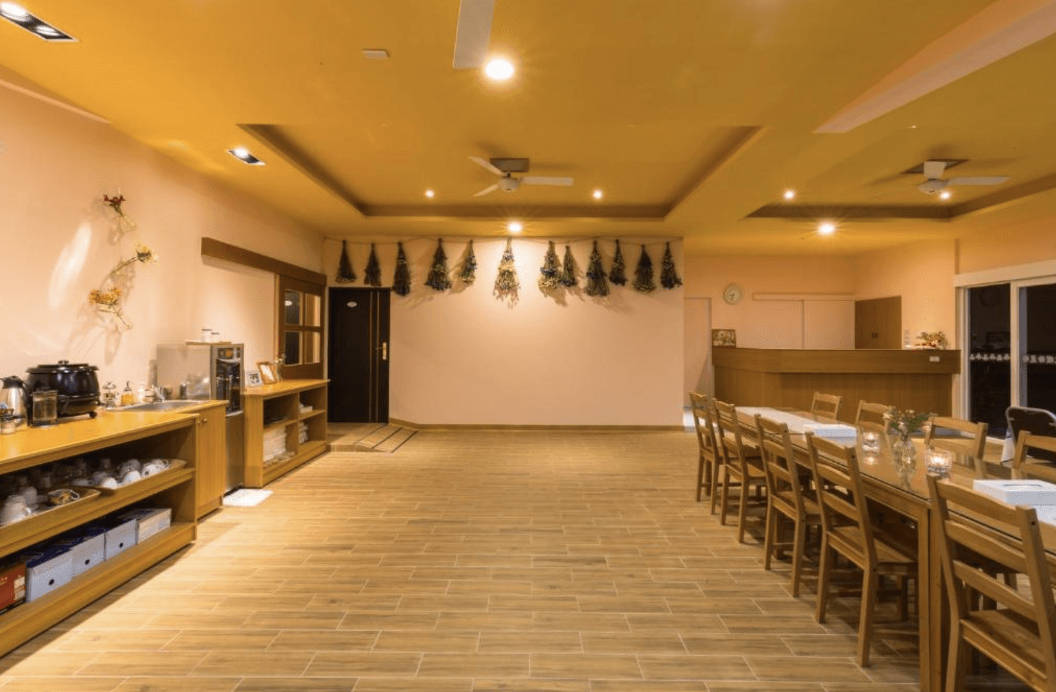 【清境農場住宿】清境繽紛農場的溫馨自然風格的用餐區與接待空間，設有長形木桌椅，牆上懸掛乾燥花裝飾，天花板配有嵌燈與吊扇，左側為餐具與飲品自助區，整體以木質元素與溫暖燈光營造出家的感覺。