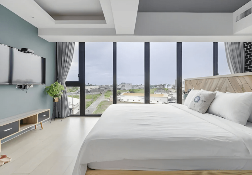 【東港住宿】"東港住宿精選示意圖"灣稻旅居的房間配有大片落地窗，可遠眺田園與城市景色，搭配柔和灰白色系牆面與簡約木質家具，床鋪設有白色床單與抱枕，營造出明亮又寧靜的舒適氛圍。