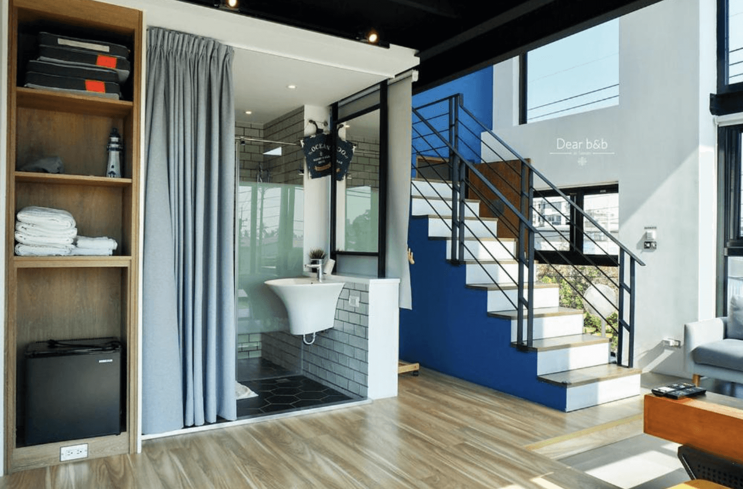 【東港住宿】灣稻旅居的挑高空間搭配開放式樓梯設計，整體以木質地板與藍白牆面為主，衛浴空間以拉簾區隔，設有洗手台與玻璃淋浴間，並配置開放式置物架與小冰箱，充滿設計感與機能性。