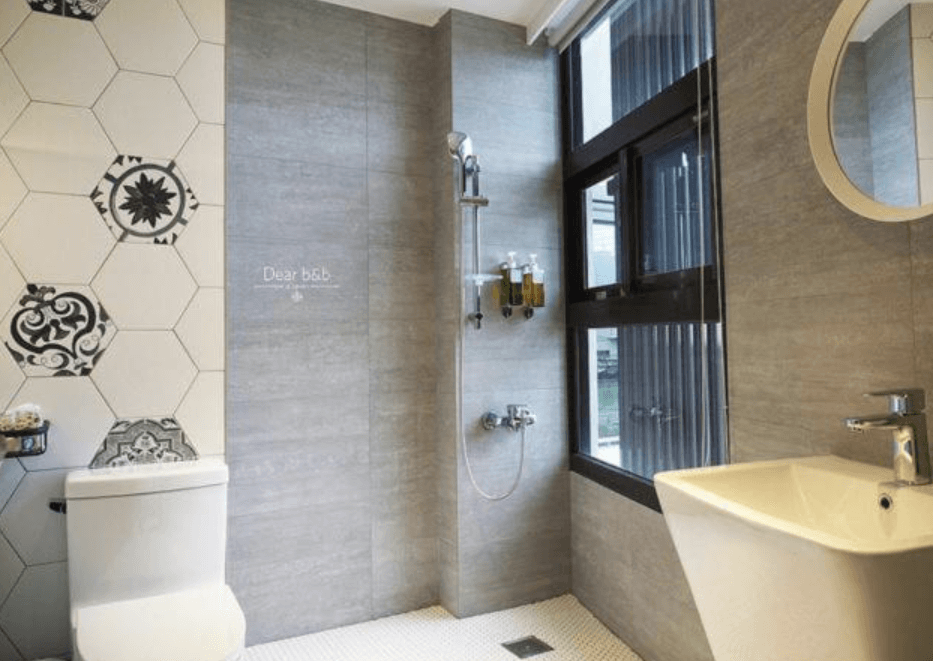 【東港住宿】灣稻旅居的浴室牆面以灰色磁磚與白色幾何花磚結合，搭配圓形鏡面與白色方形洗手台，設有淋浴設備與窗戶，自然光線充足，整體明亮整潔又具設計感。