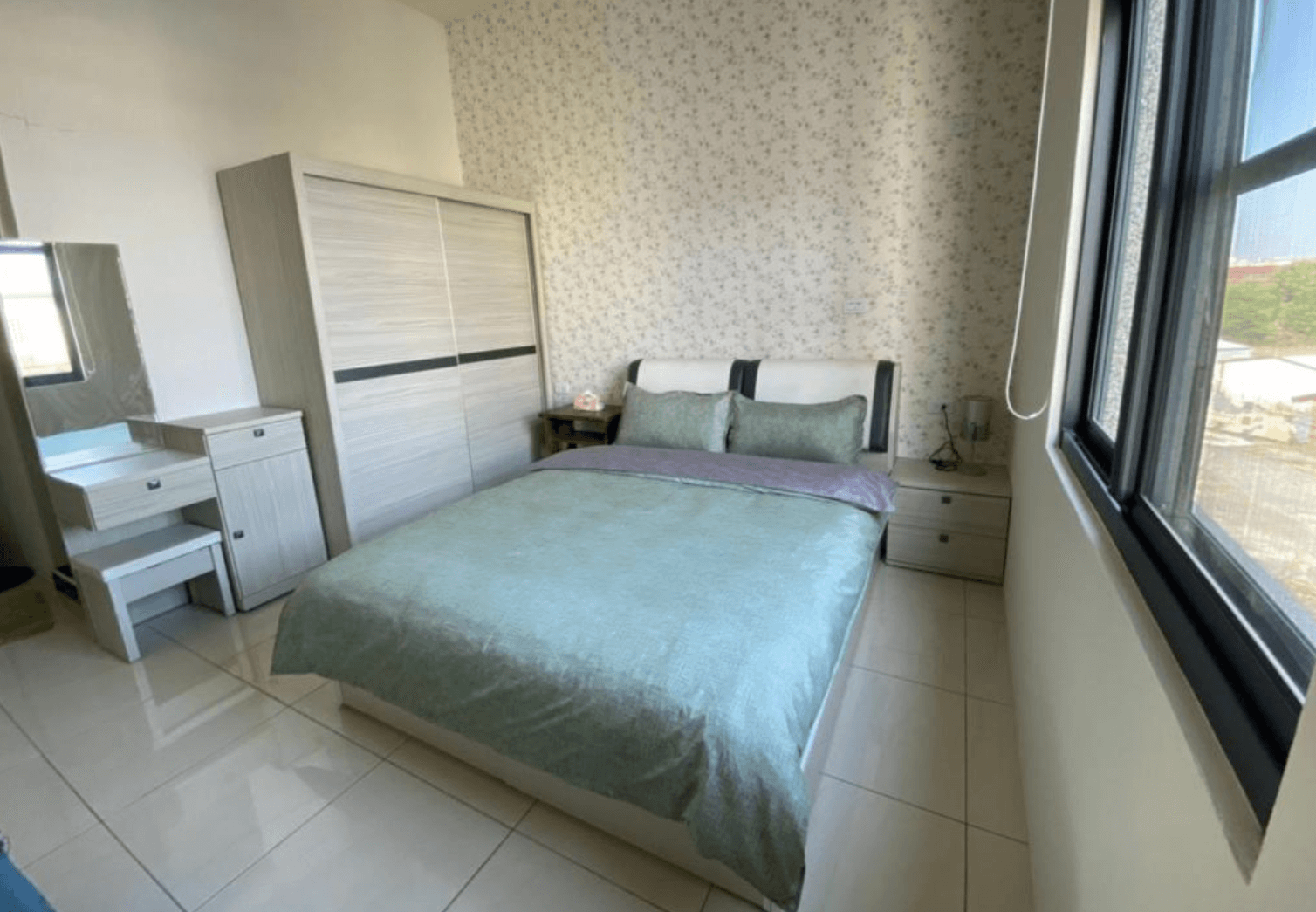 【東港住宿】留下旅舍的房間設有一張雙人床，搭配淺綠色床組與紫色被巾，牆面為碎花壁紙，窗邊採光良好，並配有衣櫃、梳妝台與床頭櫃，整體風格溫馨舒適。