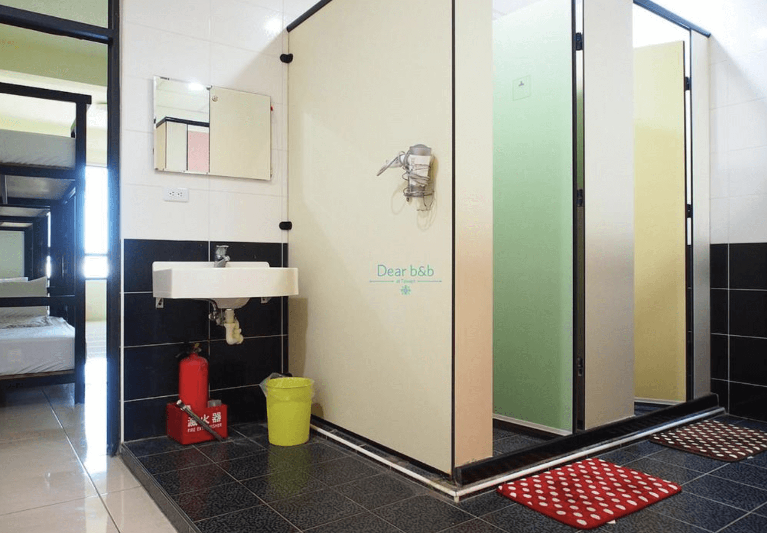 【東港住宿】留下旅舍乾濕分離設計的公共衛浴空間，設有獨立淋浴間與洗手台，搭配彩色隔間門與紅色止滑墊，牆面貼有Dear b&b 標誌，並配有滅火器與垃圾桶，維護安全與整潔。