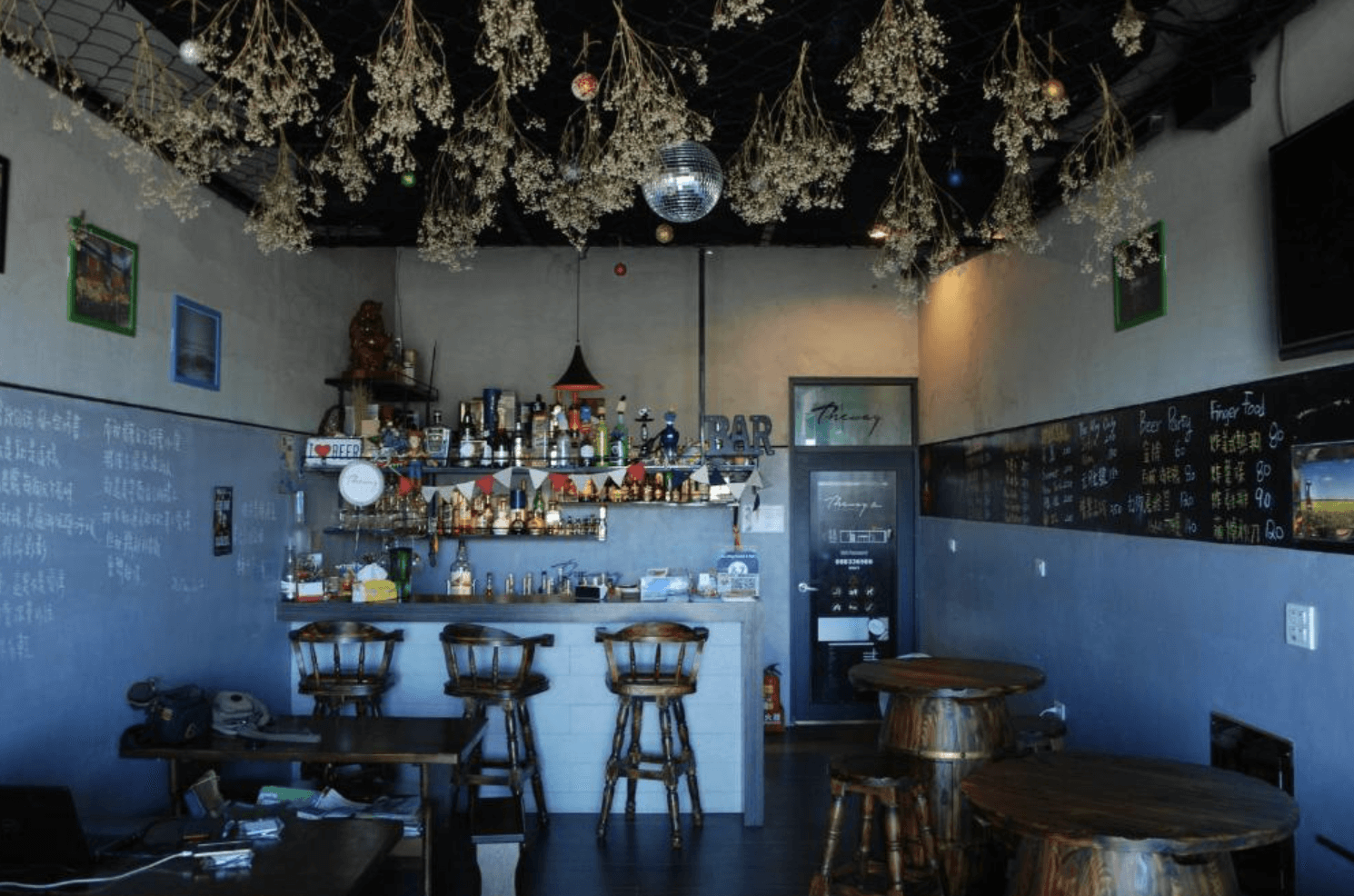 【東港住宿】留下旅舍充滿工業風格的旅舍酒吧空間，天花板掛有乾燥花與燈飾，設有木質高腳椅與圓桌，吧台擺放多樣酒品與擺設，牆上寫有手寫菜單與飲品價格，營造輕鬆熱絡的社交氛圍。
