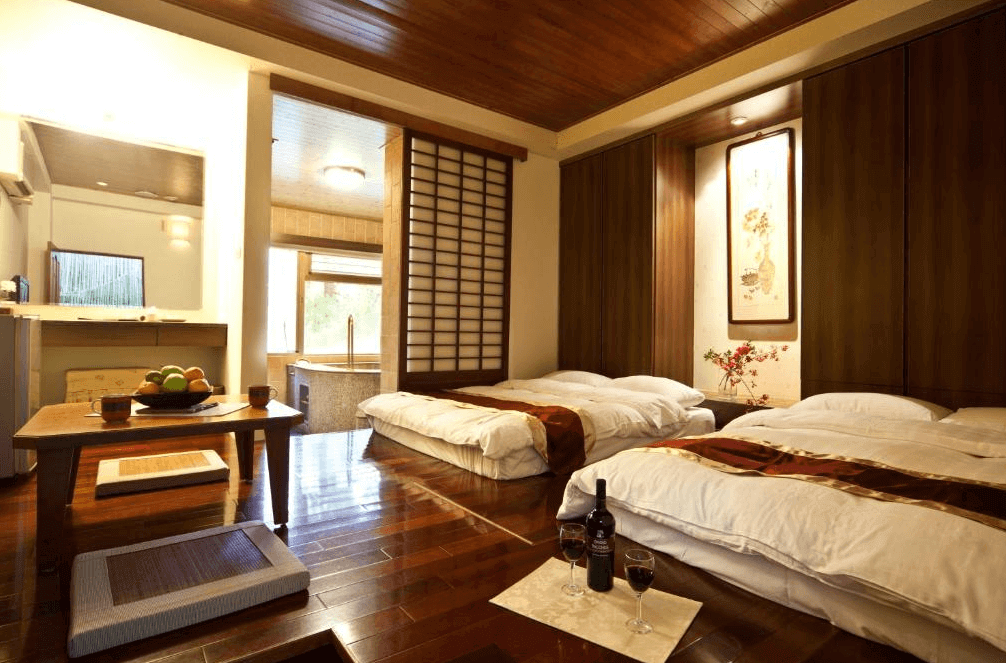 【谷關住宿】神木谷假期大飯店的房間採傳統日式風格，設有四組鋪在木地板上的厚實床墊，搭配和風拉門與掛畫，充滿東方禪意。角落配有低矮木桌與坐墊，茶具與水果盤擺設齊全，適合全家人共享悠閒時光。後方為半開放式湯屋，隱私與放鬆兼顧。