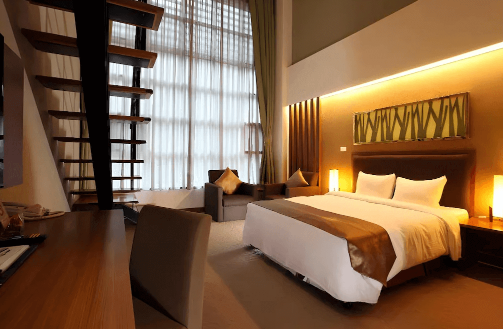 【谷關住宿】統一渡假村 谷關溫泉養生會館的房間採挑高設計，大片落地窗配上柔和窗簾，自然光灑落整個空間。木質螺旋樓梯通往樓中樓區域，房內設有一張大床、雙人沙發與書桌，整體風格溫暖沉穩，燈光柔和，營造放鬆氛圍。