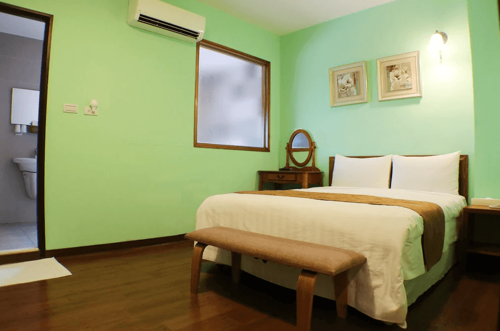【關子嶺溫泉】“白河住宿”芳谷溫泉小棧的淡綠色牆面溫馨雙人房，設有一張雙人床、木質化妝台與長凳，房內設有冷氣，浴室為獨立空間。