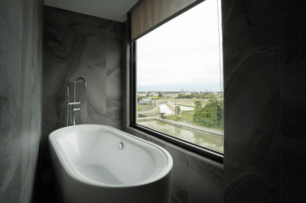 【五結住宿】藏闊渡假會館設有大型獨立浴缸的觀景浴室，搭配灰色石紋牆面與大面積窗景，可一邊泡澡一邊欣賞田園與村莊景致，營造寧靜放鬆氛圍。