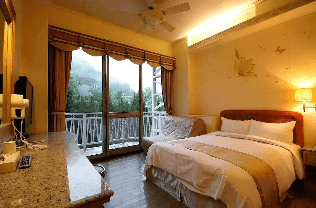 【谷關住宿】谷關 溫泉飯店的房內採柔和米黃色調搭配木質地板，設有一張舒適雙人床與小型沙發椅。大片落地窗外即為私人陽台，能眺望翠綠山景，牆面以簡約花卉與蝴蝶圖樣點綴，營造自然寧靜的休憩氛圍。