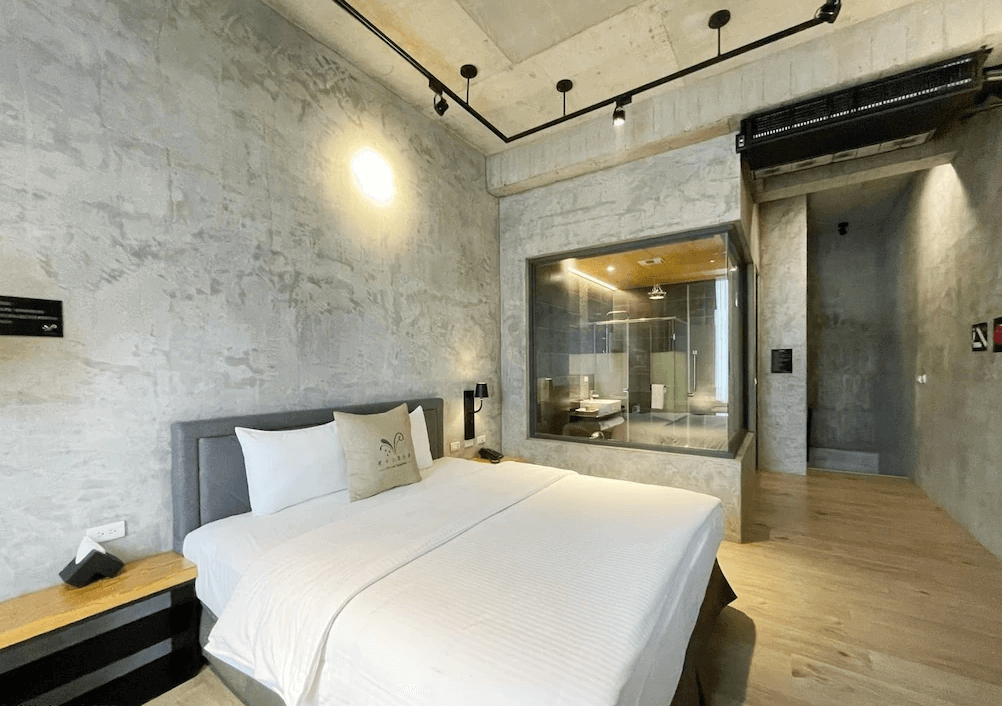 【蘇澳住宿】貳十二度行旅現代極簡風格客房，採清水模牆面與木質地板設計，設有雙人床與大片玻璃窗，內部可見衛浴空間，打造通透感十足的空間感。