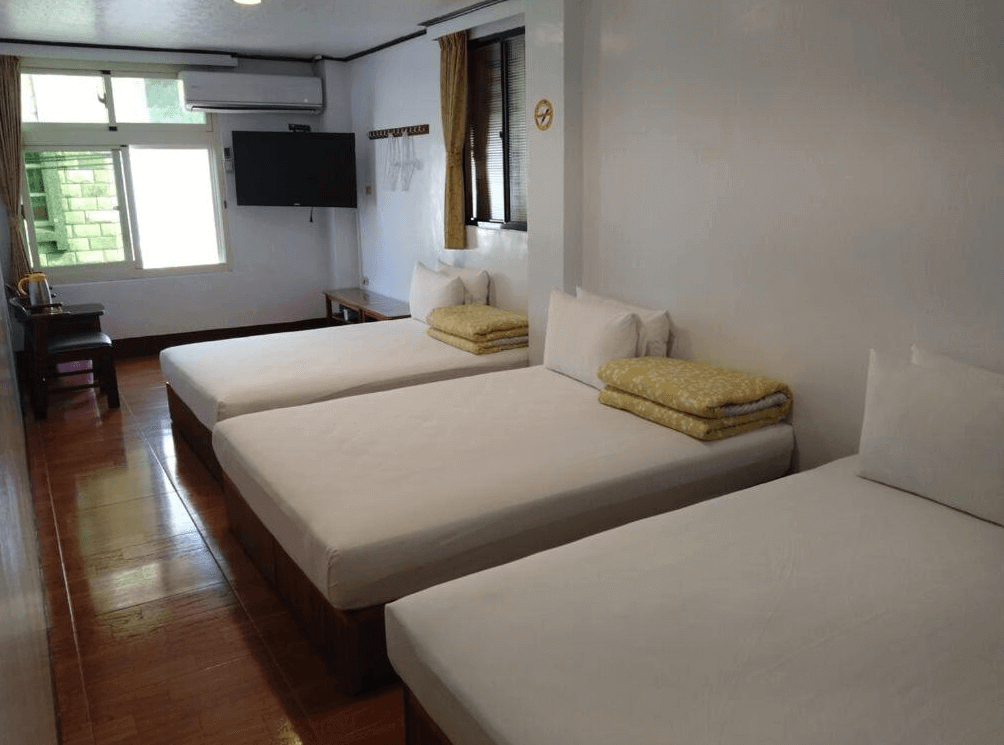 【蘇澳住宿】金華冷泉溫泉旅館的四人房空間配置三張單人床，附設電視、冷氣與窗戶，牆上掛鉤可吊掛衣物，木地板與白牆設計營造出乾淨簡單的住宿環境。