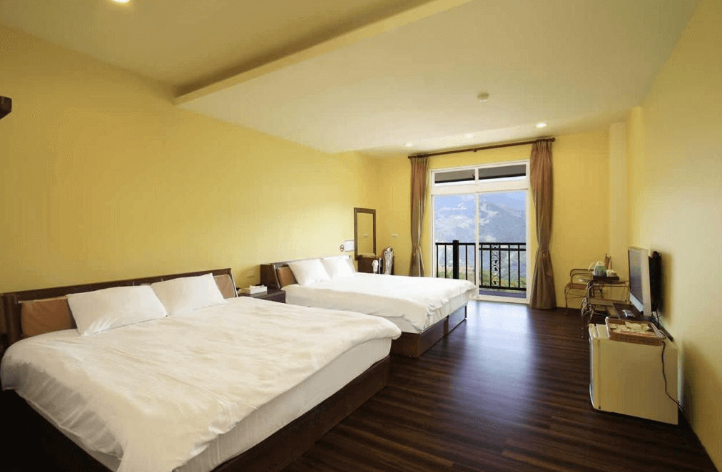 【清境農場住宿】雲濛仙境民宿的寬敞明亮的四人房，設有兩張大床，牆面以柔和黃色為主色調，搭配深色木地板，房內配有液晶電視、小冰箱與茶水區，大片落地窗外可眺望壯麗山景，空間簡約又舒適，適合家庭或朋友同住。