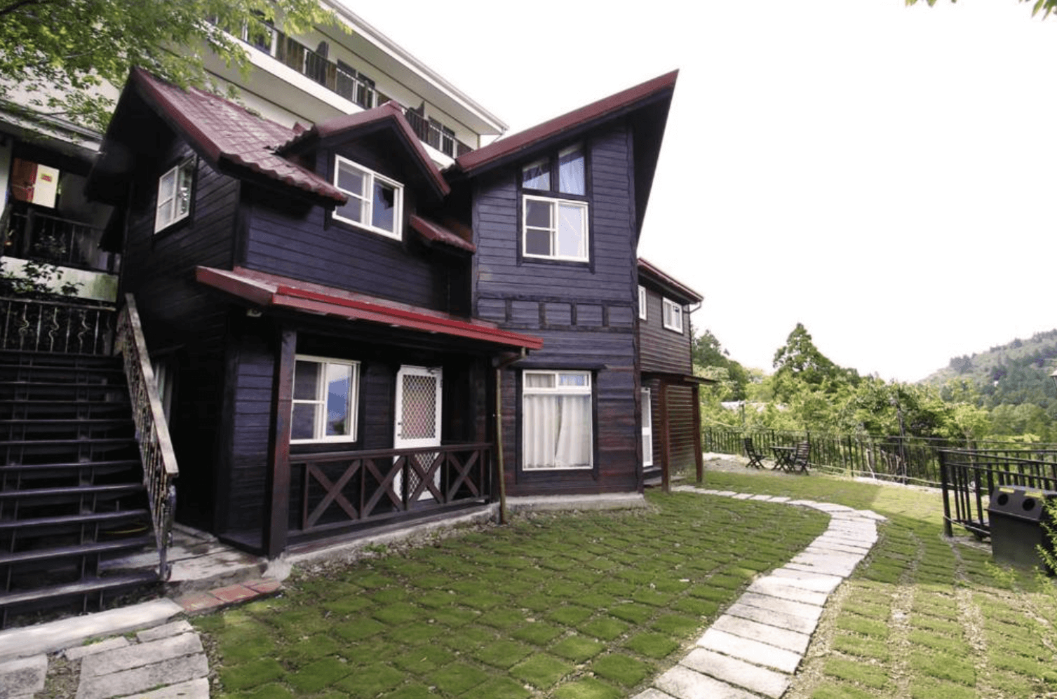 【清境農場住宿】雲濛仙境民宿的雙層深色木屋，屋頂為紅棕色斜瓦設計，前方為鋪有綠色草皮與石板步道的庭園，旁邊設有戶外桌椅可休憩，環繞著山林景致，展現靜謐自然的渡假氛圍。
