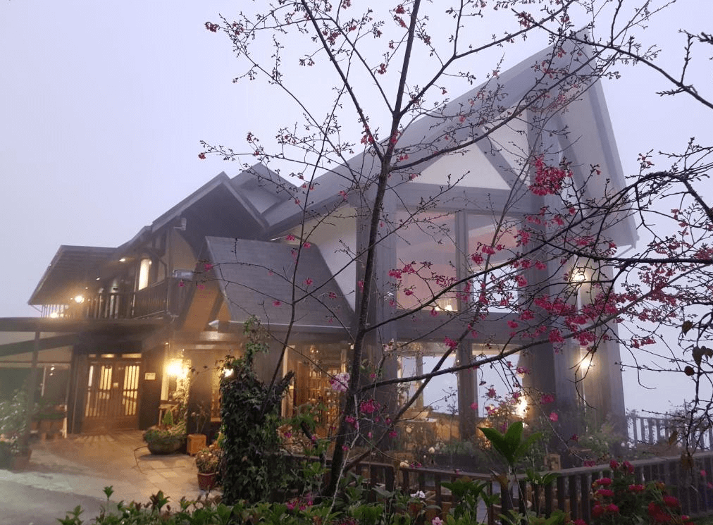 【清境農場住宿】雲頂渡假山莊雲霧繚繞的山中木屋建築，外觀採挑高斜屋頂設計，搭配櫻花樹與園藝造景，營造出夢幻又寧靜的山居氛圍，夜晚燈光柔和溫馨。