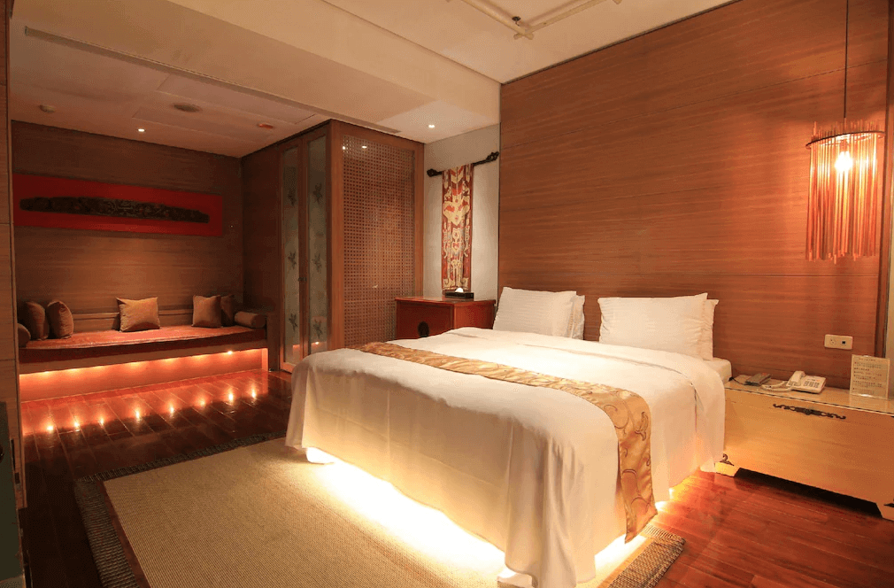 【谷關住宿】露泉渡假溫泉館的房間以原木色系為主，整體氛圍溫暖沉穩，床下與沙發區皆設有隱藏式燈光，營造柔和光影。雙人床旁擺設簡約床頭櫃與吊燈，角落還有舒適的和式沙發床區，兼具實用與美感。