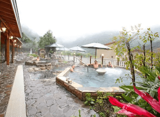 【谷關住宿】麗池山水溫泉會館的戶外溫泉區設有多個池子，石材鋪設的步道與自然植栽相映成趣。溫泉池旁擺放遮陽傘與休憩座位，山嵐環繞，空氣中瀰漫著氤氳熱氣，讓人放鬆身心、徹底享受大自然中的溫泉療癒時光。
