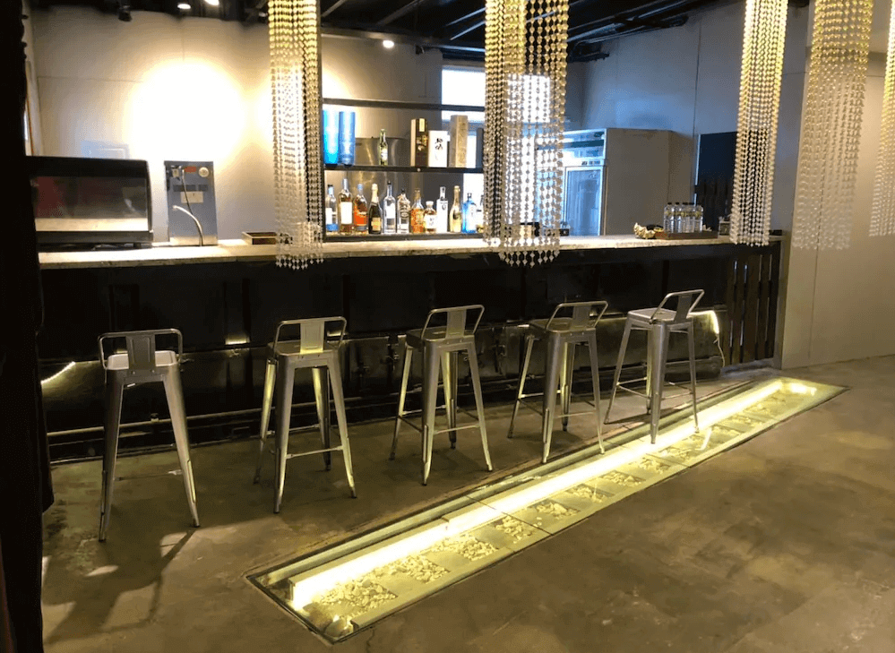 【綠島住宿】20ft 海閣的工業風格酒吧區，配有高腳椅與大理石吧檯，背後陳列各式酒類飲品，地面嵌入發光玻璃展示，氛圍時尚且具設計感。