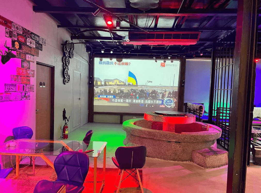 【綠島住宿】20ft 海閣的多功能交誼廳空間，搭配大型投影幕與霓虹燈光設計，中央圓形座位區結合泡腳池功能，牆上裝飾車牌與齒輪，營造出濃厚的潮流風格。