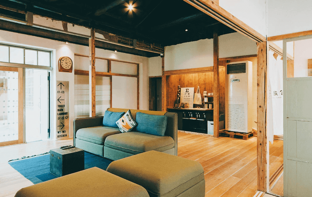 【左營住宿】Goonnight Hostel 軍旅舍公共交誼廳，溫馨舒適的交誼空間設有綠色沙發與藍色地毯，搭配原木地板與木柱結構，牆上有方向指示牌與老鐘，具文青感與機能性。