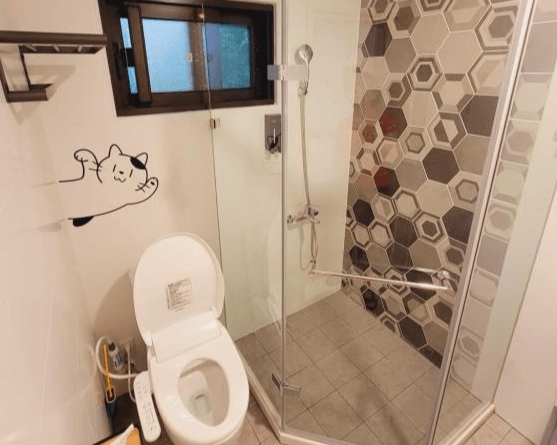 【龍潭住宿】RED ROSE 漫楓宿民宿的乾濕分離浴室，配有免治馬桶與玻璃隔間淋浴區，牆面磁磚採六角幾何設計，增添視覺層次感。馬桶旁牆面貼有可愛貓咪插畫，增添趣味與親和力，整體空間整潔實用。