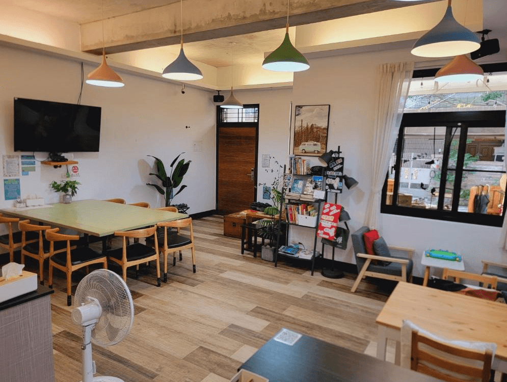 【龍潭住宿】RED ROSE 漫楓宿民宿的公共交誼廳，設有多張木質桌椅供住客使用，天花板懸掛多盞彩色吊燈，增添活潑感。牆面擺有書架與旅遊資訊，提供書籍與資訊資料。空間整潔，窗外可見在地街景，適合旅客放鬆或交流使用。