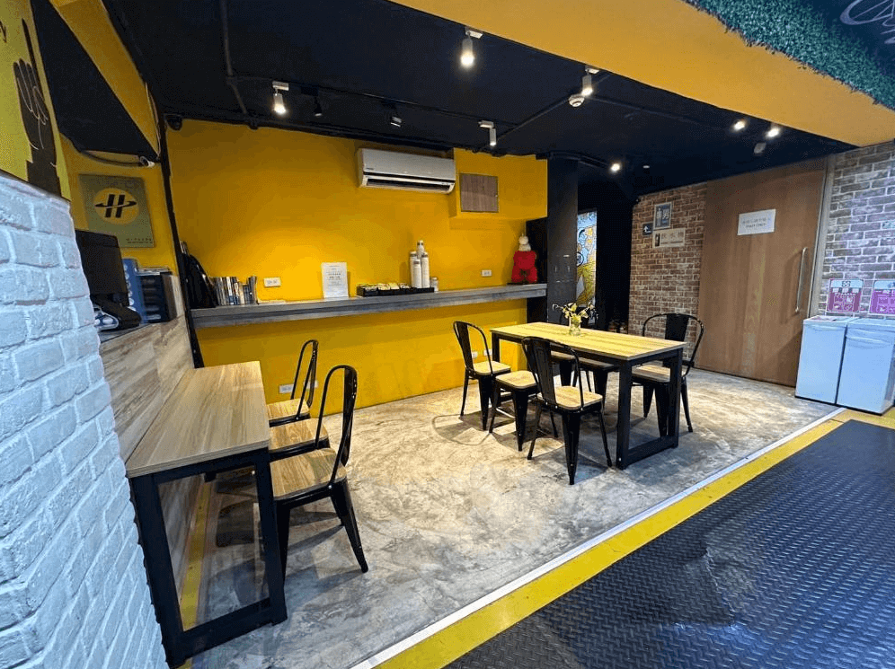 【台北車站青年旅館】Taipei Inn的交誼廳結合簡易餐飲空間，牆面為醒目的亮黃色，搭配木質桌椅與黑色金屬椅腳，氣氛活潑溫馨，適合背包客交流、用餐或短暫休息。