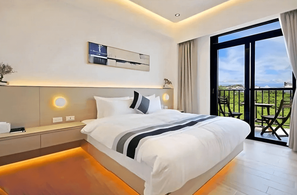 【綠島住宿】Unie會館的雙人房，配有大面落地窗與陽台，室內以柔和燈光營造溫馨氛圍，床舖整潔舒適，窗外可見綠意盎然的景致與藍天白雲。