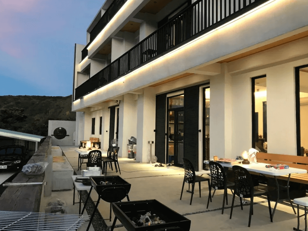 【綠島住宿】Unie會館建築外觀於傍晚時分點燈後景象，戶外空間設有烤肉區與休閒座椅，適合團體活動與親朋聚會，背景為山巒景色增添自然氛圍。