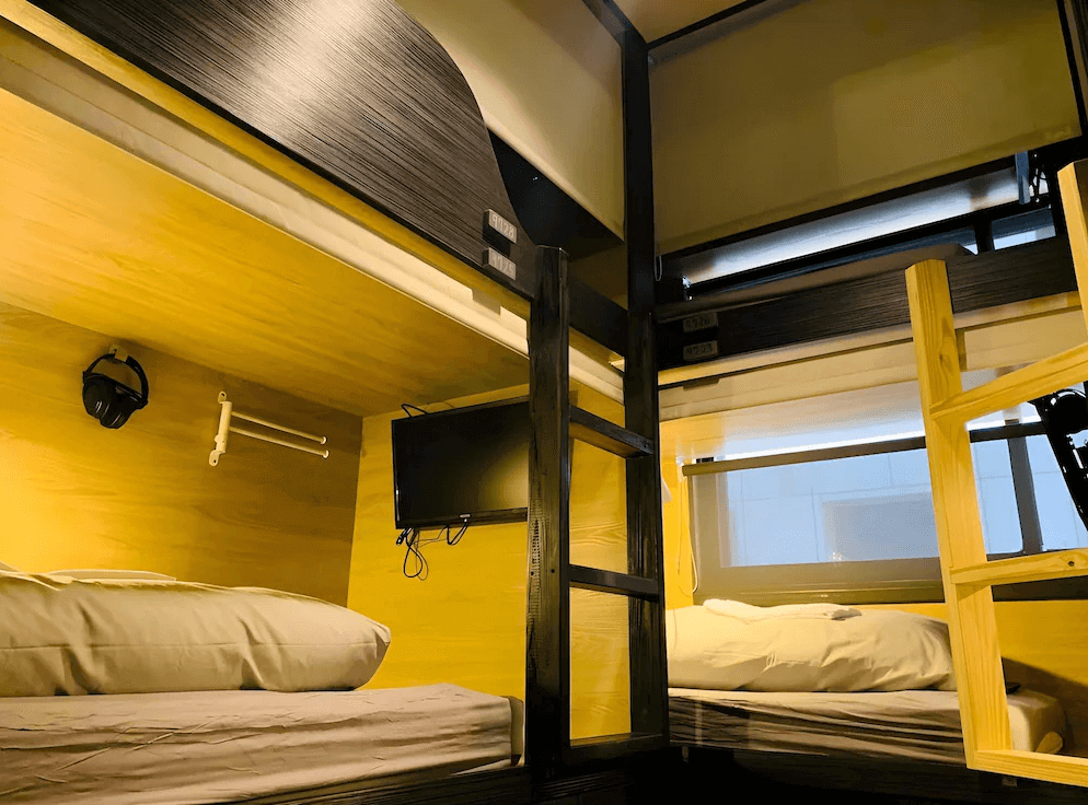 【台北車站青年旅館】WORK INN 北車館的雙層膠囊床位一隅，牆面為木質裝潢，床位配有電視、耳機掛勾與閱讀燈，整體風格溫暖且實用，兼具隱私與娛樂功能。