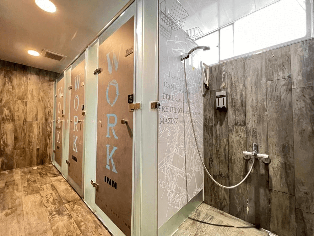 【台北車站青年旅館】WORK INN 北車館的公共淋浴間以木紋地磚搭配白色隔間門，門上印有「WORK INN」字樣，整潔明亮並設有多間獨立淋浴間與壁掛洗沐用品，營造出簡潔乾淨的洗浴空間。